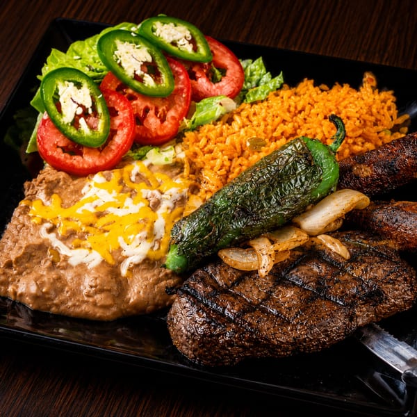 Grilled steak with refried beans, rice, salad, and a roasted poblano pepper on a black plate.