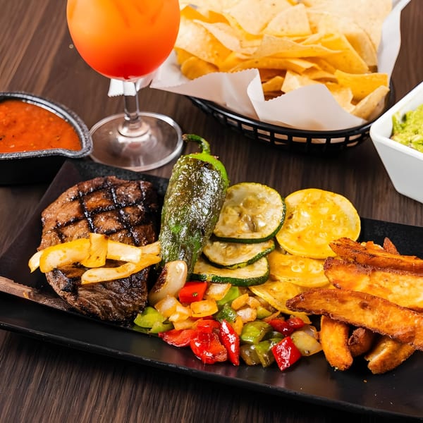 Grilled steak, vegetables, and potato wedges on a black plate with chips, salsa, guacamole, and a drink.