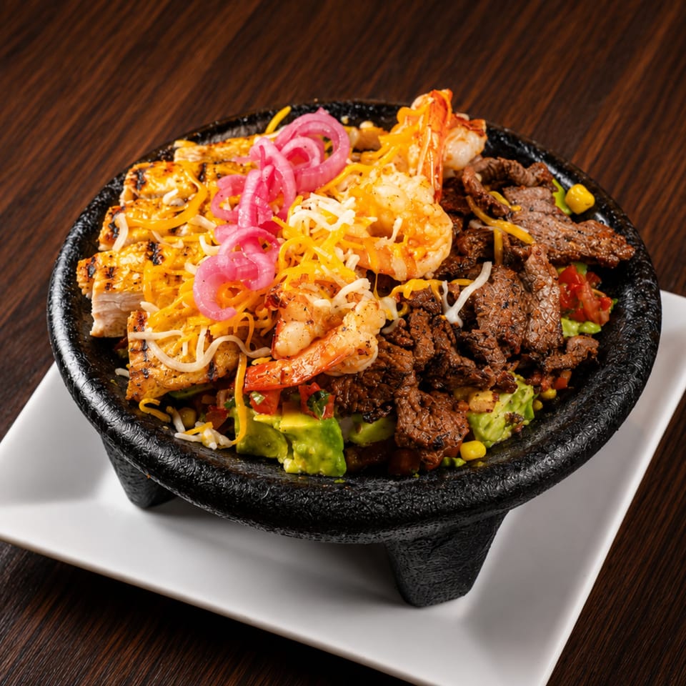 A sizzling molcajete bowl filled with grilled chicken, shrimp, steak, avocado, and pickled onions.