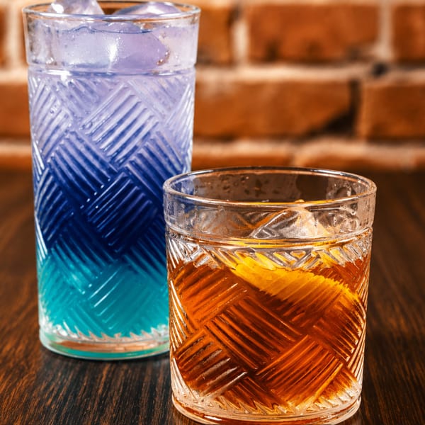 Two cocktails, one blue and one amber, in textured glasses on a wooden table.