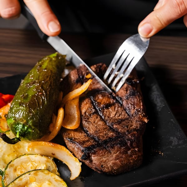 Hands cutting into a grilled steak with roasted peppers, onions, and zucchini on a black plate.
